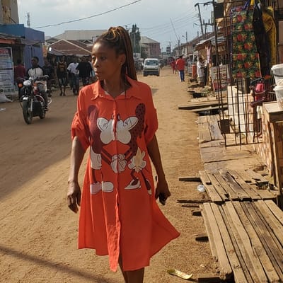 This image captures a wide-angle view of an unpaved, reddish-brown dirt street in Eziobodo, Nigeria. In the immediate foreground, a woman with light brown skin and braided hair styled in a high ponytail walks towards the viewer's left, her head turned to her left, gazing forward. She wears a knee-length, orange button-down shirt-dress featuring a prominent white graphic of Mickey and Minnie Mouse kissing. Her left hand holds a dark-colored mobile phone.
In the mid-ground on the left side of the street, a man on a black motorcycle (okada) rides towards the camera, followed by several pedestrians. The left side of the street is lined with commercial buildings, some with metal roofs; one building features a partial blue facade and a sign reading "MBG." On the right side of the street, a series of informal market stalls and shops with corrugated metal roofs extend into the background. Wooden planks are laid on the ground in front of these stalls, serving as makeshift walkways. Merchandise, including patterned fabrics, hangs from one stall, and a partial sign for "SUNSHINE Resort" is visible. Further in the background, a light-colored vehicle is observed driving away, and additional pedestrians are present. Utility poles with overhead wires are visible along both sides of the street. The sky is overcast.