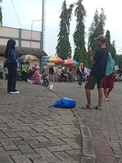 An outdoor gathering shows people near street vendors with colorful umbrellas. A blue bag lies on the patterned brick ground. A woman in a hijab stands to the left, while a man wearing a hat, t-shirt, and shorts takes a photo. Tall, thin trees are visible in the background, along with parts of a building and a tall tower. The overall scene suggests a casual, public event.