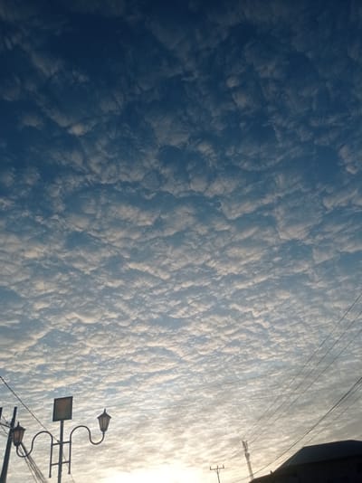 This media file is a vertical, eye-level shot of a sky filled with altocumulus clouds, creating a textured, patterned appearance. The sky is a deep blue, transitioning to a lighter, brighter hue near the horizon, suggesting it is either early morning or late afternoon. The clouds are predominantly white and gray, catching the light and casting subtle shadows, indicating fair weather.

In the foreground, silhouetted against the bright sky, are various man-made structures. To the left, there's a streetlamp with two ornate fixtures, its pole partially obscured by a cluster of dark electrical wires. A square sign is mounted on a pole above and between the streetlamp's arms. Further to the right, more electrical wires crisscross the frame. Near the bottom right corner, the dark silhouette of a building's roof is visible. In the middle distance, a cell tower stands tall, its lattice structure also silhouetted.

The scene appears to be an outdoor setting, likely in a village or town, given the presence of streetlights, utility wires, and a building. The lack of people and the natural focus on the sky suggests a moment of quiet observation or a capture of natural beauty. The overall mood is peaceful and serene, dominated by the expansive sky and its cloud formations. There is no visible text in the media file. The location context is Desa Toto Selatan, Indonesia.