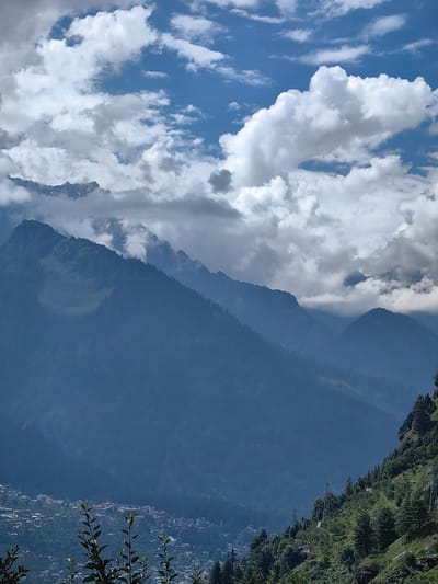 The image shows a panoramic view of a mountainous region with a town nestled in the valley below. The sky is partly cloudy with dramatic white and gray formations against a bright blue backdrop. In the foreground, lush green trees and foliage are visible, with a few branches reaching into the frame.
The midground is dominated by a series of layered mountain ranges, their slopes covered in dark green vegetation, likely forests. Some of the higher peaks are partially obscured by clouds or mist, and a few patches of snow or rocky outcrops are visible on the uppermost ridges. The mountains recede into the distance, creating a sense of depth.
In the valley, a densely populated town is spread across the lower slopes of the mountains. Buildings of various sizes and colors, including residential structures, are visible, interspersed with patches of greenery. The town appears to be built on terraced land, following the contours of the terrain.
On the right side of the image, a steep, densely forested hillside rises, with tall evergreen trees. A utility pole with multiple wires runs diagonally up this slope, suggesting the presence of electricity infrastructure. The overall scene depicts a scenic natural landscape with a human settlement integrated into the environment. The location is identified as Chhyal, India.