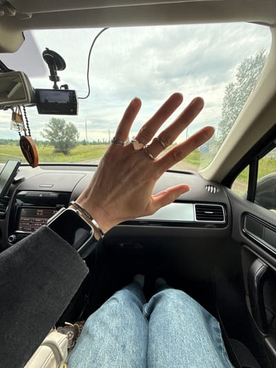 The image is a first-person perspective from inside a car, looking forward. The dashboard, steering wheel, and passenger seat are visible. A person's hand, adorned with several rings and a bracelet, is held up in front of the windshield. Through the windshield, a rural landscape with trees, fields, and a cloudy sky can be seen. A dashcam is mounted on the windshield. The scene appears to be a roadside in Zav'yalovo, Russia, as indicated by the context.