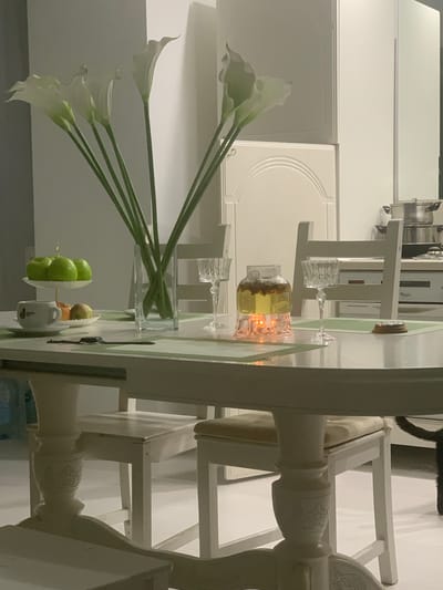 The image depicts a dining table setting in what appears to be a domestic interior, likely in Yurga, Russia. The scene is dominated by a light-colored, possibly white or cream, wooden table and chairs. On the table, a centerpiece of white calla lilies in a clear glass vase is prominent. To the right of the flowers, a decorative glass carafe filled with a golden liquid and what seems to be dried herbs or spices sits on a small illuminated base, casting a warm glow. Two crystal glasses are also visible on the table. A small bowl holding a few green apples and some other fruit is positioned further down the table. A white ceramic mug with some black markings is also visible. The overall ambiance is one of a formal or semi-formal dining arrangement, possibly set for a meal or special occasion. The lighting suggests it could be evening or an indoor setting with artificial light. The style of the furniture and the flowers evoke a sense of elegance and calm. There are no people visible in the image. The background shows glimpses of kitchen cabinets or shelving, hinting at a kitchen or dining area within a home. The time of day is not explicitly determinable, but the artificial lighting creates a cozy atmosphere.