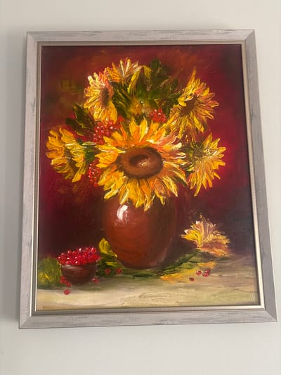 A framed oil painting depicts a still life of sunflowers in a vase, accompanied by berries. The sunflowers are predominantly yellow and orange, with thick, textured brushstrokes that emphasize their petals. Red berries are scattered throughout the arrangement, some in a small, dark bowl to the left of the vase, and others loose on the surface below. The vase is a deep brown, with a glossy finish. The background is a rich, dark red, transitioning to lighter tones in some areas, creating depth. A single fallen sunflower head with yellow petals rests on the surface to the right of the vase. The painting is framed in a light gray, textured wooden frame. The artwork is hung on a white wall. The provided context mentions the city of Pereiaslavske, Ukraine, but the image itself does not contain any visual cues to confirm this specific location.
