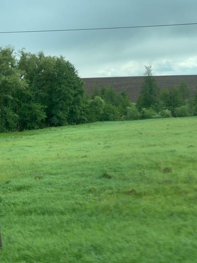 The image depicts a landscape near Kokino, Russia, featuring a green field and forested area under an overcast sky. A large, wall-like structure with a dark, earthen color is visible behind the trees. A power line runs along the left side of the frame. There is no indication of people, actions, or interactions.