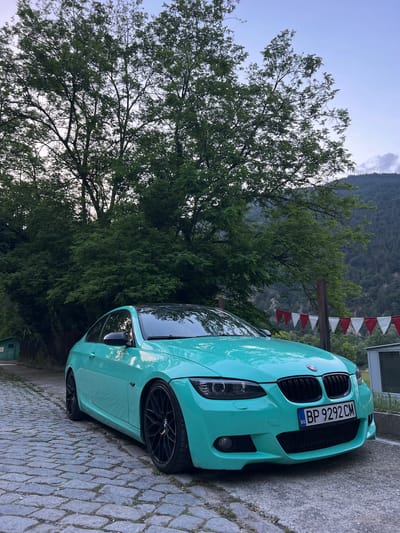 A turquoise-colored BMW 3 Series coupe (E92) with black multi-spoke wheels, a black roof, and a black kidney grille is parked in Bachkovo, Bulgaria. The vehicle's front is angled slightly right, resting on a cobblestone surface on its left side and a darker, possibly asphalt, pavement on its right. A Bulgarian license plate, "BP 9292 CM," is affixed to the front bumper. Behind the car, a dense canopy of large green deciduous trees fills the midground, with steep, heavily forested hills rising in the distant background under a light blue sky dotted with scattered white and grey clouds. To the far left, a small, weathered light green wooden shed is partially visible behind a fence. On the right side of the frame, a string of alternating red and white triangular pennants is strung between two wooden posts, with a white rectangular structure visible behind them. No people are present in the image.