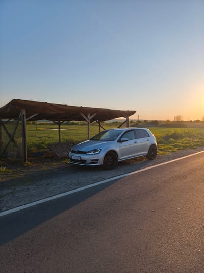 A silver Volkswagen Golf sits parked off to the side of a road in Trastikovo, Bulgaria. The car is positioned next to a makeshift shelter constructed of wooden supports and a draped fabric roof. In the background, a field stretches towards the horizon under a gradient sky, transitioning from blue above to a warm sunset glow on the right. A thin white line delineates the edge of the road, which takes up the foreground of the image. The setting appears rural, with limited signs of urbanization visible. The license plate number on the car is A 0127 PH.