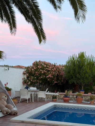The image shows an outdoor patio area with a swimming pool and lush vegetation. The sky is a soft pink and blue, suggesting either sunrise or sunset. Palm fronds hang into the frame from the top left and top right, framing the scene. In the foreground, a portion of a blue swimming pool with a decorative tiled edge is visible. To the left, a large, plush teddy bear sits on a white cushioned lounge chair under a canopy. Behind the lounge chair, a white wall with a dark security camera is visible, along with some terracotta pots containing plants. Further back, a white outdoor dining set with chairs surrounds a round table. A dense bush with abundant pink flowers dominates the middle ground, situated between the white wall and a tall, spiky green tree. Several potted plants, including cacti and succulents, are arranged along a stone wall that separates the patio from the rear vegetation. The overall impression is a tranquil and well-maintained outdoor living space. The specific location of Mont-roig del Camp, Spain, is not explicitly evident from visual cues within the image itself.