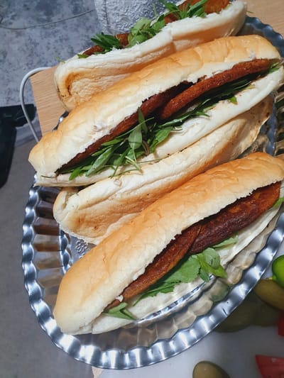 A serving platter of sandwiches, possibly a local delicacy from Kheyrabad, Iran, is presented. The platter is ornate, made of a reflective metal with a wavy edge. It contains at least three sandwiches, each served on a long, light-colored bun. The sandwiches appear to be filled with a dark, elongated piece of meat, likely a kebab or fried cutlet, and a generous portion of fresh green leafy vegetables, possibly arugula or parsley. One sandwich is visible at the top of the platter, and two more are stacked below it, with the closest one in sharp focus. The lighting suggests an indoor setting, perhaps a kitchen or dining area, with a slightly textured grey surface visible in the background and a wooden board beneath part of the platter. Additional accompaniments, such as pickles and tomato slices, are partially visible on the right side of the image.