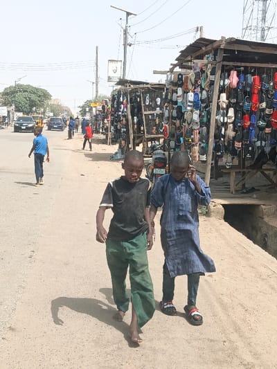 The image captures an active street scene in Hotoro South, Nigeria, under clear daylight conditions. In the foreground, two young males walk leftward on a dirt shoulder adjacent to an asphalt road. The boy on the left wears a black t-shirt with white trim and green pants, appearing barefoot. The boy on the right wears a blue plaid tunic over dark pants and black Adidas slide sandals; he holds a dark object to his right ear, consistent with a mobile phone interaction.
To the immediate right, a series of makeshift market stalls line the dirt shoulder. These structures are built with wooden frames and corrugated metal roofs and are densely packed with footwear, predominantly colorful sandals and slides, displayed by hanging. A motorcycle, bearing Nigerian license plate "JGE 671VF," is partially visible parked near these stalls. A concrete drainage ditch runs along the right side of the road.
On the asphalt road, a young male in a blue shirt and dark pants walks away from the camera. Further down the street, another individual in a red shirt walks towards the right. Several vehicles, including a yellow taxi and a black sedan, are present on the road. Multiple utility poles, some with overhead wires, extend into the background; one features a solar panel at its apex. A tree with green foliage is visible on the left side of the road in the mid-distance. Shadows are cast to the left, indicating a sunlit environment.
