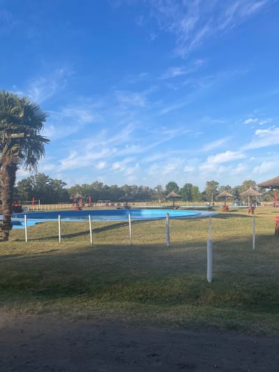 A landscape shot displays a blue pool surrounded by grass, with evenly spaced white vertical poles lining one side. Beyond the pool, trees and thatched umbrellas are visible. To the left, a clear blue sky features wispy clouds, and the top of a palm tree is visible in the lower left corner. A dirt path appears on the right side of the grassy area.