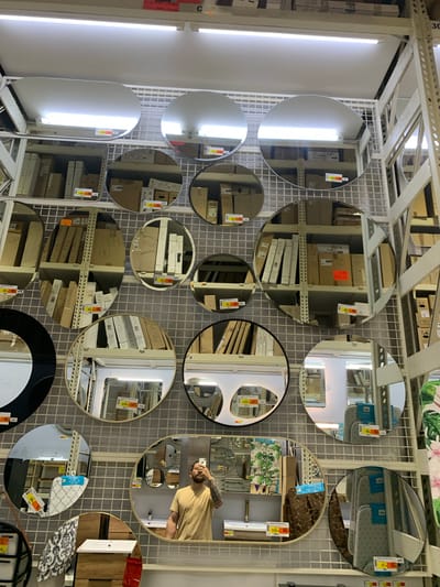 The image shows a retail display of various mirrors on white grid shelving. Multiple mirrors of different shapes and sizes are hung, reflecting the surrounding environment. The reflections reveal aisles with more merchandise, including boxes and shelving units. Some mirrors have price tags visible. In the lower right quadrant of the image, a man is visible in a mirror, holding up his phone and taking a selfie. He is wearing a tan t-shirt and has tattoos on his arm. The man is in a store, likely a home improvement or department store, given the shelving and merchandise. The lighting appears to be artificial, with fluorescent lights visible. The overall scene is a retail display aimed at showcasing mirrors for sale. The location is Berģi, Latvia.