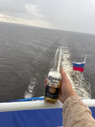 The image shows a person's hand holding a nearly empty bottle of Corona Extra beer. The bottle is clear glass with a yellow label featuring the brand name "Corona Extra" and the slogan "LA CERVEZA MAS FINA." The label also indicates an alcohol content of 4.5% and that it is a beer. The background suggests a body of water, likely a river or lake, with choppy waves and a wake behind it. In the distance, a small flag with red, white, and blue stripes, possibly the Russian flag, is visible on a pole. The overall lighting suggests it is daytime, and the presence of water droplets on the bottle and railing, along with the somewhat overcast sky, indicate potentially rainy or misty weather. The setting appears to be outdoors, on what seems to be a boat or a dock, with a blue railing visible in the foreground. The mood could be interpreted as relaxed or celebratory, given the presence of a beer and the scenic outdoor environment. The location context provided is Belous, Russia.