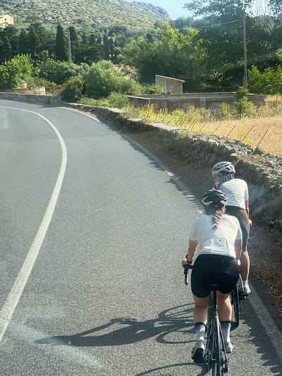 Two cyclists, one in front of the other, ride along a paved road in Pollença, Spain. Both are wearing white cycling jerseys, dark shorts, helmets, and cycling shoes. The road has a white line marking the edge of the lane. The surrounding environment features a dry, grassy embankment with a stone wall, and lush green trees and vegetation on a hillside in the background. The sky is clear and bright. The image appears to be taken from a slightly elevated perspective, possibly from another vehicle following the cyclists.