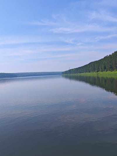 The image shows a wide, calm river with a forest lining the far bank. The sky is blue with scattered white clouds. A transmission tower is visible on the left side of the forest. The scene is likely in the city of Piskunovka, Russia, given the mention of its location in the prompt. There are no people or animals visible in the image. The water reflects the trees and sky, creating a serene natural landscape.