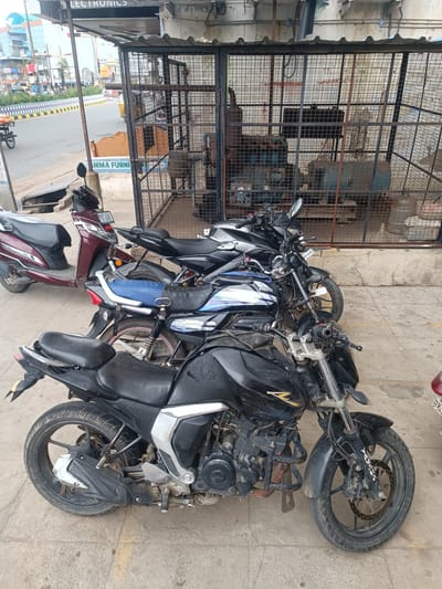 This media file captures an outdoor scene in Kurnool, India, likely during the daytime under clear weather conditions. The foreground is dominated by several parked motorcycles and a scooter. On the left, a maroon scooter is prominently displayed, with its license plate "AP40A 1896" clearly visible. To its right, a black motorcycle with a blue seat is parked, followed by another black motorcycle in the immediate foreground. The motorcycles are lined up on a concrete surface, suggesting a street or a parking area.
Behind the vehicles, a caged area encloses some industrial-looking equipment, possibly machinery or generators. A portion of a building facade is visible above, with some signage. One sign appears to read "MMA FURNI," suggesting a furniture shop nearby. Another sign above it reads "LECTRONICS." The overall setting seems to be a commercial area with a mix of shops and public spaces. The presence of multiple parked vehicles indicates a potentially busy area. The lighting suggests it is daytime, with no strong shadows indicating it might be late morning or early afternoon.