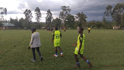 The image depicts a grassy field in Bungoma, Kenya, located at the coordinates 0.554 latitude and 34.774 longitude. The field is surrounded by trees and there is a small building in the background. A group of people are playing soccer on the field. Two men in the foreground are facing away from the camera, while two men in the center are facing the camera. The men in the center are wearing bright yellow shirts with black trim and black shorts. The men in the foreground are wearing casual clothes, and the man on the left is wearing a gray sweater. One man is walking to the right and the other man is walking to the left. The grass is green and there are clouds in the sky. The trees in the background are all different shapes and sizes.