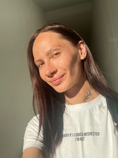 A young woman with long, dark hair is captured in a close-up, outdoor portrait. She is wearing a white t-shirt with the text "SORRY IF I LOOKED INTERESTED I'M NOT" printed on it. The woman has several piercings, including a nose ring and multiple earrings. A tattoo of a floral vine is visible on her neck. She is illuminated by natural sunlight, which casts shadows across her face and the background. The environment appears to be a street or walkway in the city of Pervomaysky, Russia, with a muted, grey wall as the backdrop.