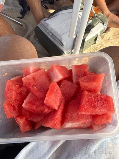 The image shows a clear plastic container filled with cut pieces of watermelon. The watermelon pieces are vibrant red and appear to be fresh, with visible seeds in some pieces. The container is placed on a textured surface that appears to be sand, suggesting an outdoor setting. In the background, a person's legs are visible, one with shorts and the other with a tanned leg. A backpack and a beach chair are also present, indicating a beach or picnic setting. The lighting suggests it is daytime, likely sunny. This scene is consistent with a relaxed day at the beach, potentially in a location like Costa da Caparica, Portugal, known for its expansive beaches.