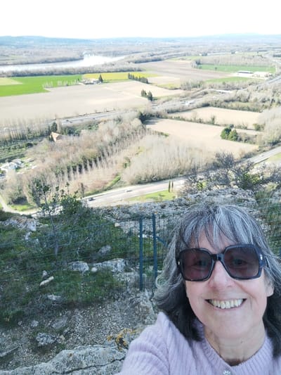 A happy woman takes a selfie from a high vantage point, overlooking a vast and scenic landscape. She is positioned in the lower right portion of the frame, smiling broadly at the camera. She wears large, dark, rectangular sunglasses with thick frames and a light pink, possibly fuzzy or knitted, long-sleeved top with a small, sparkly embellishment on the left side of her chest. Her hair is medium length, a mix of gray and darker strands, and slightly tousled, framing her face.
Behind her, a green wire mesh fence stretches across the middle ground, indicating a viewing area or barrier. The immediate ground around her is rocky and uneven, with sparse green vegetation and moss.
The background unfolds into a wide, expansive valley or plain, viewed from above. Several roads crisscross the landscape. A prominent multi-lane highway or dual carriageway runs horizontally across the mid-distance, with a few cars visible on it, appearing as small white and dark specks. Another smaller road winds through the lower left, leading towards a cluster of buildings that resemble a small village or residential area. An elevated road or bridge crosses over the main highway further to the right.
The land is primarily composed of agricultural fields. Many fields are light brown or tan, suggesting they are barren or recently tilled. Interspersed are patches of vibrant green fields, likely showing early spring crops, and in the far distance to the left, a field with bright yellow sections, possibly indicating blooming rapeseed or similar.
A wide river or large body of water snakes across the upper left portion of the landscape, its banks lined with dense vegetation, mostly bare trees. Numerous trees, predominantly deciduous and without leaves, line the roads, fields, and riverbanks, giving the impression of late winter or early spring. Rows of tall, slender trees, possibly cypresses or poplars, are visible along some field edges. A few scattered buildings, including a farmhouse with a reddish-brown roof, are nestled within the fields. Distant hills or low mountains are faintly visible on the hazy horizon.
The sky is bright and clear, indicating a sunny daytime scene. The overall impression is one of tranquility and beauty, captured by the woman's joyful expression as she enjoys the panoramic view.