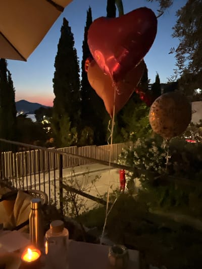 This is a vertical, dimly lit nighttime photo taken outdoors. The subjects are primarily balloons, with one heart-shaped red balloon prominent in the foreground, appearing to be tethered by a thin white string. Another round, patterned balloon is visible in the midground, also tethered. The scene suggests a festive or celebratory occasion.
The setting appears to be a natural outdoor environment with trees and foliage in the background, suggesting a garden, park, or backyard. In the midground, there is a railing or fence, possibly part of a balcony or patio. Beyond that, a dimly lit road or pathway is visible, with some illuminated windows in buildings further in the distance. The sky is a deep blue, transitioning to lighter hues at the horizon, indicating it's either dusk or dawn. There are hints of mountainous terrain and distant lights, suggesting a scenic location, potentially near a body of water. The presence of a lit candle or lantern in the bottom right corner further adds to the nighttime atmosphere, casting a warm glow.
The overall mood conveyed is somewhat whimsical due to the balloons, but the low light and outdoor setting at night create a sense of intimacy or quiet celebration. There is no visible text in the image.