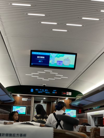 The image shows the interior of a train carriage, likely on a high-speed rail line in China, specifically noted as Longhua District. The scene is oriented vertically, suggesting the photo was taken with the camera held in portrait mode.
The primary subjects are several passengers, including a young girl with dark hair and a blue hair accessory, seated and looking towards a screen. Another passenger with dark hair, wearing a striped top and a white collared shirt, is partially visible, also looking towards a screen. The train carriage is characterized by a modern, clean design with ribbed white walls and integrated LED lighting strips.
Several digital displays are visible. One prominent screen shows a cartoon character, likely a game or entertainment interface, with Chinese text. Another screen displays train information, including the date "2026-06-26", time "16:37", the current station "前方到站" (approaching station), and the next station "虎门" (Humen). A larger display panel on the right side shows the train number "8" and indicates "武汉,下一站:虎门" (Wuhan, next stop: Humen), along with interior and exterior temperatures: "内温25℃" (indoor temperature 25°C) and "外温30℃" (outdoor temperature 30°C). Another visible text reads "ZES 325900 定员49", which might be related to the train's model or capacity.
A red emergency button and some other warning signs are also present on the wall. The overall atmosphere appears calm, with passengers engaged with their screens or resting. The time indicated is late afternoon, and the exterior temperature suggests a warm day. The text "臺奶倾倒后方茶杯" (Milk to be poured behind the teacup) is visible on a white surface, possibly a warning or instruction related to food and drinks.