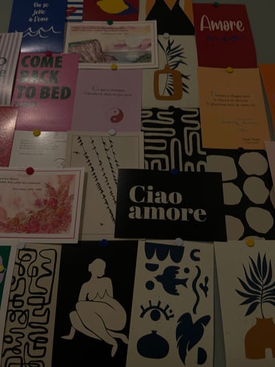 The image shows a collection of cards pinned to a wall, creating a mood board. The cards feature various designs and texts, including the phrase "Amore mio" in red, "Ciao Amore" in black and white, "Come Back to Bed" in pink, and "On se jette à l'eau" in blue. Other cards have inspirational quotes in French, signed by Mark Twain and Victor Hugo. There are cards displaying nature scenes, abstract shapes, and an illustration of a nude figure. Pins of different colors secure the cards in place.