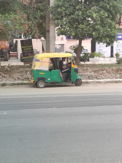 A green and yellow auto-rickshaw is parked on the side of a road in Gurgaon, India. The auto-rickshaw has advertisements on its sides. A man is seated inside the auto-rickshaw, looking at his phone. In the background, there are trees, buildings, and street signs. One sign advertises "Prime Salon Academy - Unisex Salon" with a phone number. Another sign appears to be for SBI (State Bank of India). The environment suggests an urban or semi-urban setting with road infrastructure and commercial establishments.