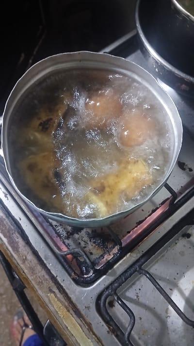 A pot is simmering on a gas stove. The pot contains water and what appears to be food. The stove is dirty and stained. Part of a person's leg, wearing a blue top and sandals, is visible in the bottom right corner. The photograph was taken in the city of Putucual, Venezuela.