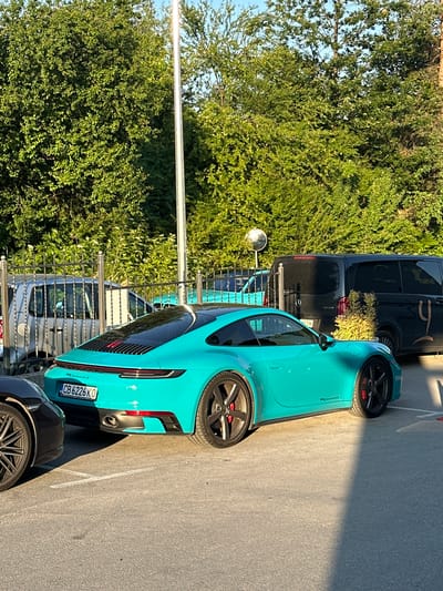 A turquoise Porsche 911 Carrera S is parked on asphalt in front of a fence and lush green trees. The car's license plate reads "CB 6226 KO". Behind the Porsche, a black Mercedes-Benz Vito van is visible, with a stylized gold logo on its side. A silver car is parked behind the fence, and a portion of a dark-colored car is visible in the foreground. A tall, white pole stands near the trees. The environment suggests an outdoor setting, possibly a hotel parking lot or a similar establishment. The lighting indicates it is daytime, with shadows cast by the cars and the pole. The location is identified as Kerenite, Bulgaria.