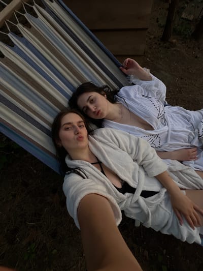 Two young women are lying side-by-side in a striped hammock. The woman in the foreground is closer to the camera and has her lips pursed. Both women are wearing white robes. The hammock is suspended outdoors, with dry grass and dirt visible below. In the background, there are trees and wooden structures. The lighting suggests it is either early morning or late evening.
