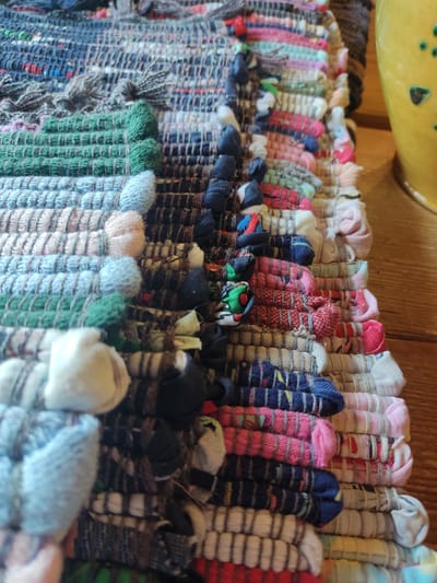 The image presents a detailed, high-angle perspective of a hand-woven, multi-colored rag rug, resting on a light-colored wooden surface. The rug is constructed from distinct horizontal bands of fabric strips in various hues, including prominent sections of green, light blue, pale pink, white, dark blue, red, and diverse mixed patterns, creating a textured and somewhat irregular surface. Visible individual fabric wefts and knotted ends are apparent along its edges, indicative of its artisanal craftsmanship. To the right of the rug, a partial view of a yellow ceramic object, adorned with green circular designs, is visible. The underlying wooden surface displays discernible grain patterns. No individuals are present in the scene, and no specific actions are depicted. While the prompt indicates a location in the city of Staraya Russa, Russia, the visual content of the image provides no specific identifying cues to confirm this geographic location.