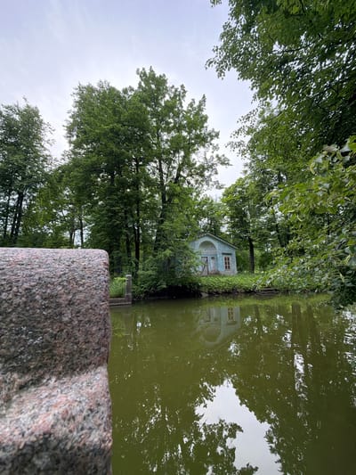 The image depicts a serene scene in Pushkin, Russia, likely during the daytime under an overcast sky. A large, textured granite boulder dominates the foreground, partially obscuring a tranquil body of green water. The water reflects the lush, verdant trees that line its banks, creating a mirrored effect.
Nestled amongst the foliage, a small, light blue pavilion with a distinctive arched facade and two windows is visible. Its reflection is clearly visible in the still water, suggesting a peaceful and undisturbed environment. The surrounding vegetation is dense and green, with various trees and bushes contributing to the natural beauty of the setting. The overcast weather contributes to a soft, diffused light, enhancing the tranquil mood of the scene. No people or text are visible in the image.