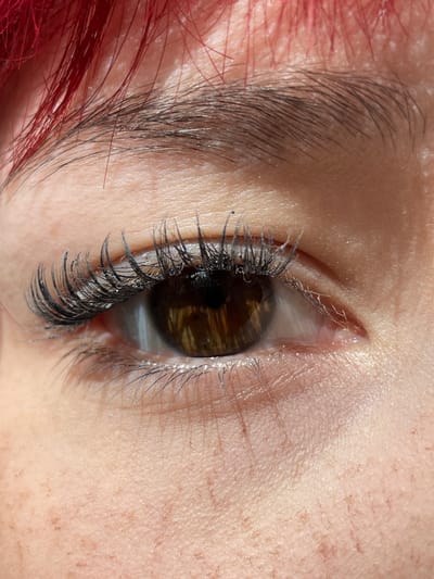 The image is a close-up, high-angle shot of a human eye, focusing on the eyelashes, iris, and surrounding skin. The subject's hair, which appears to be dyed bright red, is visible at the top of the frame. The eyelashes are long and appear to be enhanced with mascara, with some clumping visible. The iris is a rich brown color with subtle golden flecks. The skin around the eye shows texture and some light freckling. The lighting suggests natural light, possibly sunlight, creating highlights and shadows. There is no indication of the city of Солнечная Поляна, Russia, in this image.
