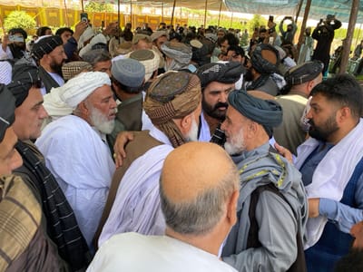 The image shows a crowd of men gathered in an outdoor setting in Zaker-E Sharif, Afghanistan. The men are predominantly wearing traditional Afghan clothing, with many wearing white turbans and long white robes. Some are wearing dark blue or black turbans and robes. Several men are dressed in more modern attire, including a blue checkered shirt and a light blue long-sleeved shirt. Some men are wearing traditional woven shawls or scarves. Many men are embracing or shaking hands, with a general sense of camaraderie and warmth in their interactions. The background shows a mix of greenery and structures, suggesting a rural or urban setting with a mixture of modern and traditional elements. The environment appears to be sunny and clear. There are no women present in the image.  The location of the scene, based on the provided coordinates, is in the Zaker-E Sharif district of Balkh province, Afghanistan.