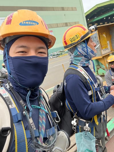 The image shows two construction workers in Geoje-si, South Korea, wearing orange hard hats and safety harnesses. The worker in the foreground is a man wearing a blue face mask and a blue beanie. He has a blue and yellow safety harness with a rope and safety glasses hanging from it. He is looking directly at the camera. The second worker, to the right, is also wearing an orange hard hat, a blue long-sleeved shirt, and a safety harness. He has a headlamp attached to his hard hat and is smoking a cigarette. He is looking to his right. The background shows a large industrial building with green siding and a yellow structure with Korean writing on it. The ground appears to be paved.