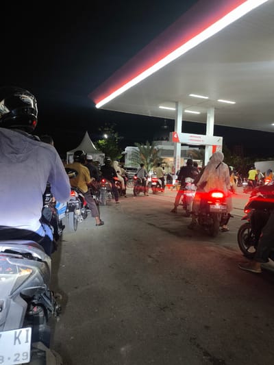 A nighttime scene at a Pertamina gas station in Lhoksukon, Indonesia, depicts a long queue of motorcycles and their riders waiting to refuel. The gas station canopy is illuminated by bright white lights and edged with red trim, casting a glow on the scene below. Several motorcycles are parked side-by-side, with riders seated on them. Some riders are wearing helmets, while others have them on their heads or are not wearing them. The ground is asphalt, and the overall atmosphere suggests a busy, operational gas station at night. In the background, a white tent structure is visible.
