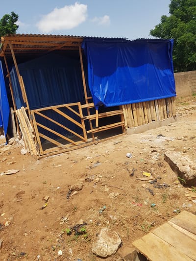 This media file depicts an outdoor scene in Palade, Nigeria, possibly a construction site or a makeshift shelter. The main subject is a partially constructed structure covered with a bright blue tarp. The structure appears to be made of wood and possibly corrugated metal or plastic sheeting for the walls and roof. A wooden door frame with diagonal wooden slats is visible, suggesting an entrance. Below the blue tarp, some corrugated blue material can be seen, which might be part of the building's walls.

The ground around the structure is a mix of dry, sandy soil with scattered debris. This debris includes pieces of plastic, paper, and other discarded items, indicating an area that may not be regularly cleaned. Some sparse dry grass and a few small green plants are growing through the soil. In the background, a large green tree with dense foliage is visible on the left side of the frame, and a brick wall can be seen on the right. A large, light-colored rock or concrete chunk is also present in the foreground on the right.

The sky is visible in the upper left portion of the image, with a few white clouds against a clear blue backdrop. The lighting suggests it is daytime, likely late morning or early afternoon, with bright sunlight casting shadows. No people are visible in the image. The overall impression is one of a functional, possibly temporary, structure in a somewhat undeveloped or unkempt environment. There is no visible text in the image.