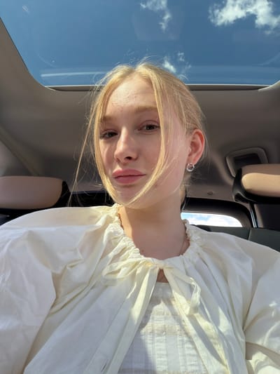 This media file is a portrait of a young woman with blonde hair, taken from a slightly angled, close-up perspective. She is wearing a white, voluminous blouse with gathering at the neckline. Her left ear is visible, adorned with a small, circular silver earring. Her skin is fair and appears illuminated by natural light, casting subtle shadows on her face. Her gaze is directed towards the camera.
The background suggests an indoor setting, likely a car, with hints of car interior elements such as a headrest and a dark seat. A significant portion of the image is dominated by the sky, visible through what appears to be a sunroof or window. The sky is bright blue with scattered white clouds, indicating it is daytime with clear weather.
The overall mood appears casual and relaxed, possibly a selfie taken during a car ride. The lighting suggests it is either morning or afternoon, given the brightness of the sky. No text is visible in the image. The provided location context is Rossiyskiy, Russia.