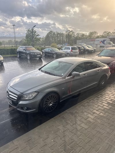 The image captures a rainy, overcast scene in a parking area or alongside a road, likely during morning or late afternoon given the diffused light. Multiple cars are parked, their surfaces glistening with water droplets from ongoing or recent rainfall.
In the foreground, a grey Mercedes-Benz sedan is prominently featured, parked on a wet, dark paved surface next to a sidewalk made of interlocking concrete blocks. Its front grille and partial license plate, which reads "HH 7731", are visible, suggesting a German registration. To its left, partially obscured by the Mercedes, is a dark Audi SUV or car with a visible partial license plate "E 553HI", hinting at Spanish registration, despite the stated location of Kraishte, Bulgaria. Further down the row, a white hatchback (possibly a Dacia Sandero) and other vehicles are parked.
In the middle ground, a building with a "Pireco" logo is visible, flanked by trees with varying densities of foliage. The background reveals an expanse of cloudy sky, with some brighter patches suggesting the sun's presence. In the far distance, snow-capped mountains rise above the horizon, partially veiled by mist or clouds, indicating a mountainous setting. A tall metal power line tower and streetlights punctuate the landscape. The overall atmosphere is quiet and somewhat muted by the rainy weather.