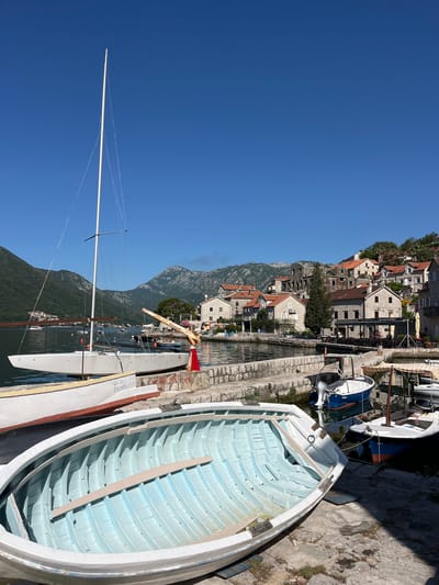 The image depicts a serene waterside scene in Perast, Montenegro. In the foreground, a white rowboat with a light blue interior is resting on a stone embankment, angled towards the viewer. Behind it, a larger white sailboat, named "Aurora Pearl," is moored, its tall mast reaching into the clear blue sky. To the right, several smaller boats are docked, including a blue and white motorboat and a sailboat with a striped canopy.
The background reveals the charming town of Perast, characterized by its stone buildings with terracotta roofs, nestled on a hillside overlooking the water. The Boka Kotorska bay stretches out, with lush green mountains forming a dramatic backdrop. The water is calm, reflecting the clear sky and the surrounding architecture. The scene is bathed in bright sunlight, indicating a pleasant day. There are no visible people in the image, and the overall impression is one of tranquility and picturesque coastal life.
