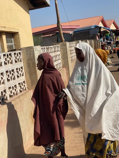 The image displays an outdoor scene in Samaru, Nigeria, under bright daylight. In the foreground, two women walk from left to right. The woman on the left, positioned in profile facing left with her mouth slightly open, wears a dark reddish-brown head covering and a loose matching garment over patterned trousers. She holds the right forearm of the woman to her right. This second woman, facing slightly towards the viewer, wears a white head covering and a loose white garment featuring a circular green emblem inscribed with "SAMARU BRANCH." She also wears patterned trousers.
To the immediate left, a low wall constructed from patterned concrete blocks runs parallel to a light yellow-ochre building with a single window. In the mid-ground, a tall wooden pole stands vertically. The background reveals a bustling market environment with multiple stalls situated under structures topped with orange-red corrugated metal roofs. Various goods are displayed on the stalls, and several other individuals, including one person in a yellow garment bending over a stall, are visible further back. The ground appears to be unpaved dirt, and strong sunlight casts distinct shadows across the scene.
