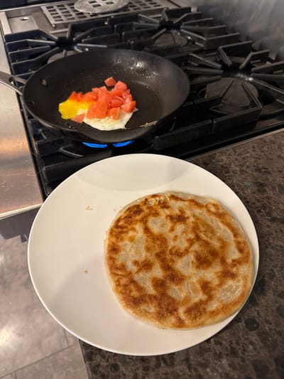 A forensic description of the scene in Cresskill, United States:

The image depicts a cooking scene within a kitchen. In the midground, a black frying pan rests on a professional-grade gas stovetop with black cast-iron grates. Inside the pan, one partially cooked egg with a distinct bright yellow yolk and set white is accompanied by a pile of diced red tomatoes. A blue flame is visible beneath the pan, indicating an active gas burner. To the right, on a dark, mottled countertop, a white, round ceramic plate holds a single, golden-brown, circular flatbread with a textured, layered surface, consistent with a paratha. A section of light-colored tiled flooring is visible in the lower left corner. No individuals are present in the frame.