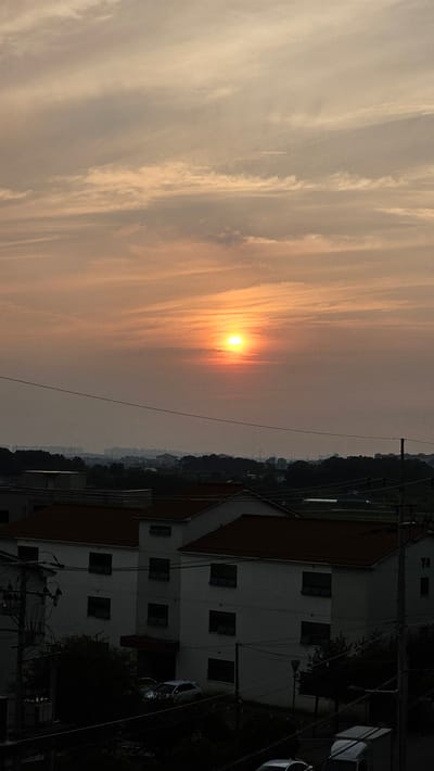 The image captures a sunset over Asan-si, South Korea. The sun, a bright orange orb, is low in the sky, casting a warm glow through hazy clouds. In the foreground, the rooftops of white buildings with red-tiled roofs are visible, suggesting a residential area. Several utility poles and wires are also present, indicating the presence of infrastructure. The landscape beyond the buildings is dotted with trees and hints of distant structures. The overall scene depicts a tranquil evening in a suburban or semi-urban setting.