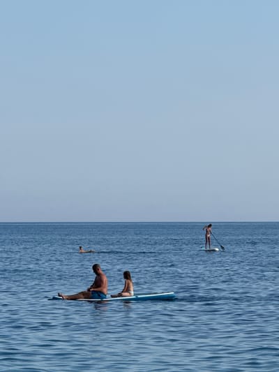 The image depicts a serene scene of people paddleboarding on a calm, blue body of water under a clear, pale blue sky. The water is rippled, suggesting a gentle breeze. The perspective is from above, looking down at an angle, with the water occupying the right half of the frame and the sky the left.
There are three individuals visible, all on paddleboards. One person is further in the distance, standing upright on a paddleboard and holding a paddle. Closer to the foreground, two other individuals are engaged in activity on their paddleboards. One, wearing blue shorts, is bent over, seemingly adjusting something or preparing to dive. Another person, wearing a light-colored bikini bottom, is also bent over on their paddleboard. A paddle is visible near the first person. There are also a few small, detached objects floating in the water.
The overall atmosphere is one of leisure and outdoor recreation on a bright day. The clear sky suggests fair weather, likely daytime. The repetition of the blue water and sky creates a peaceful and expansive feeling. No visible text is present in the image. The location context provided is Mirište, Montenegro.