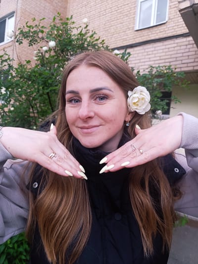 A fair-skinned woman with auburn hair stands outdoors, posing with her hands near her face. She has a white rose tucked behind her ear. The woman wears rings on her fingers. She is dressed in a black jacket and a grey sweater, and a dark button can be seen on her jacket. The background includes a brick building and green foliage. The location is Chernogolovka, Russia, based on the filename of the image.
