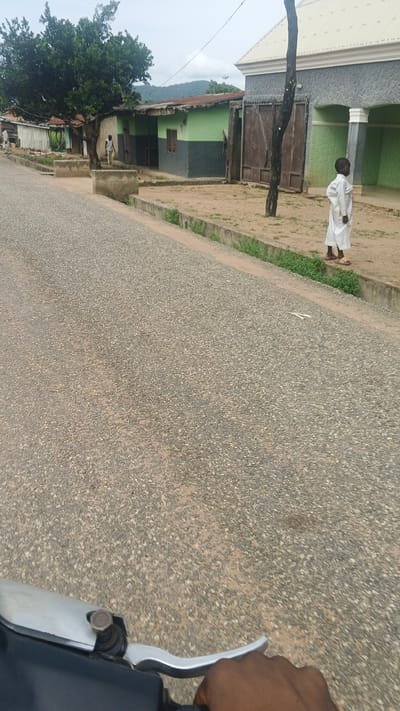 This is a first-person perspective video of someone riding a motorcycle on a paved road in Serti, Nigeria. The road is bordered by a dirt path and a concrete curb with sparse green vegetation. On the right side of the road, there are buildings with green and gray walls. A young boy in a white robe stands near one of the buildings. A large tree with green foliage stands to the left of the buildings. In the background, rolling hills are visible under a cloudy sky. The overall atmosphere appears to be peaceful and rural. The time of day is likely daytime, given the lighting. There is no visible text in the image.