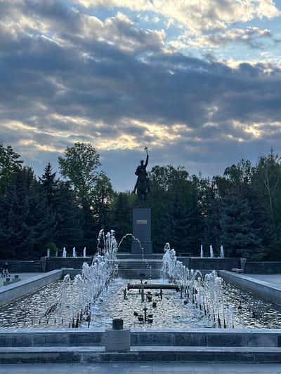 The image, correctly oriented, captures a majestic outdoor scene featuring a prominent fountain and a historical monument. In the foreground, a long, multi-tiered fountain made of dark stone or concrete showcases multiple jets of water actively spraying, creating a shimmering effect on the water's surface as it reflects light.

Behind the extensive fountain, a dark-colored statue stands tall on a rectangular pedestal, partially obscured by trees. The statue appears to depict a heroic figure, possibly on horseback or in a powerful pose, holding a raised spear or standard. Faint text, likely "MIHAI VITEAZUL," is visible on a plaque on the statue's base, identifying it as a monument to Michael the Brave.

A dense treeline, comprising both coniferous and deciduous trees, forms a natural backdrop behind the monument and fountain. Above the trees, the sky is partially overcast with dramatic, textured clouds, allowing patches of bright sunlight to break through, suggesting either late afternoon or early evening. A small, distant cross is faintly visible against the sky above the treeline.

The overall scene conveys a sense of grandeur and tranquility. A very faint human figure, possibly with a bicycle, can be glimpsed near the far edge of the fountain structure. The setting appears to be a public square or park, consistent with the provided location context of Craiova, Romania.