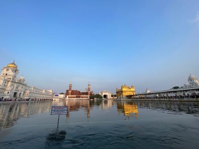 The image depicts the Golden Temple in Amritsar, India. In the foreground, the calm water of the Amrit Sarovar (holy pool) reflects the surrounding architecture. A sign in Punjabi and English stands in the water, stating "Please do not throw eatables in holy pool." In the mid-ground, the iconic Golden Temple complex is visible. The main shrine, known as Harmandir Sahib, is a golden edifice surrounded by water. To its left, a white marble building with domes and minarets stretches along the water's edge, and to the right, a long colonnade is lined with people. A red brick building with multiple domes and tall minarets is also present in the scene, along with a white archway. The sky above is a clear, pale blue, suggesting it is either early morning or late afternoon. People can be seen walking along the walkways of the temple complex and sitting by the water. The overall scene is serene and majestic.
