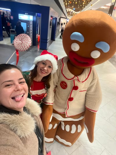 Three people pose together, including a gingerbread man mascot. Two women are in the foreground, one sticking out her tongue. The second woman wears a Santa hat and a red and green elf-like costume. The gingerbread man is in the back, behind the two women. The background includes a blue and silver bank of machines and a blurry image of people walking. A large lollipop is to the left of the woman in the Santa hat.