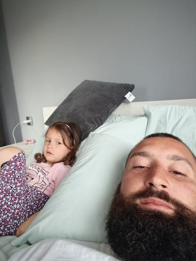 A selfie is taken from a low angle, featuring a man with a full beard and a young girl lying in bed. The man's face, partially obscured by his beard, fills the right side of the frame. The young girl, with brown hair and wearing a pink t-shirt with a graphic design, is positioned on the left, propped up against a pillow. She has a pink headband and patterned pajama bottoms. The background shows a light blue duvet and a grey pillow with a tag attached. A white wall with a power outlet and a charging cable is visible behind the girl. The scene appears to be indoors, likely a bedroom. The lighting suggests it is daytime. The girl's expression is somewhat serious, while the man's face is more neutral. No specific text is clearly legible besides the design on the girl's shirt and the tag on the pillow, which is too blurry to read. The location is identified as Sandanski, Bulgaria.